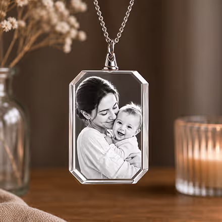 Rektangulärt 2D-kristallhalsband graverat med ett porträtt av en mamma och bebis, upphängt i en silverkedja bredvid ett tänt ljus.