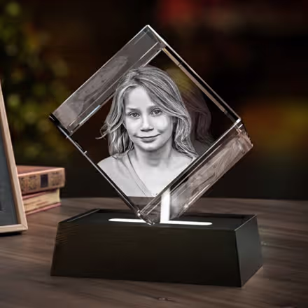 Diamantformad 3D-fotokristall graverad med en leende ung flicka, placerad på en svart LED-ljusbas bredvid originalfotot.