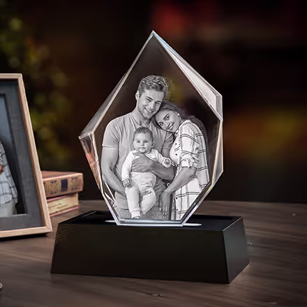 Isbergsformad 3D-fotokristall graverad med ett porträtt av föräldrar och deras bebis, placerad på en svart LED-ljusbas bredvid det ursprungliga familjefotot.